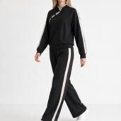 Model wearing a black tracksuit with white side stripes on the sleeves and pants, paired with white sneakers.