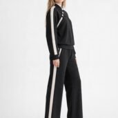 Model wearing a black tracksuit with white side stripes, standing in profile with white sneakers exposing a casual athletic look.