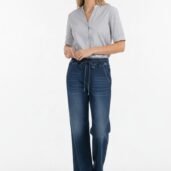 Woman in a light gray button-down blouse paired with dark wash wide-leg jeans with a drawstring, standing in a studio setting.