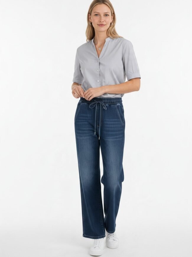 Woman in a light gray button-down blouse paired with dark wash wide-leg jeans with a drawstring, standing in a studio setting.